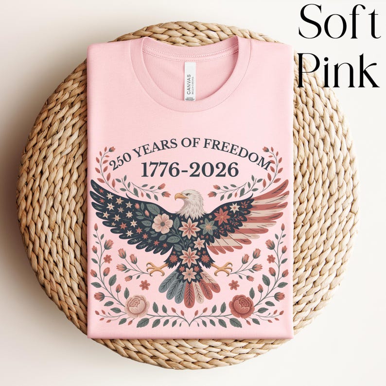May include: A soft pink t-shirt with a round neck featuring a patriotic design. The graphic includes an eagle with floral accents, the American flag, and the text "250 YEARS OF FREEDOM 1776-2026". The shirt is displayed on a woven, circular surface.