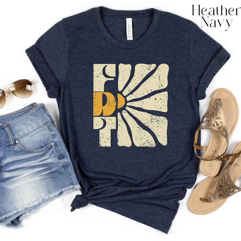 May include: A heather navy blue t-shirt featuring a vintage-style graphic of a sun with the word "FRI" in a retro font. The graphic is in shades of yellow, orange, and white.