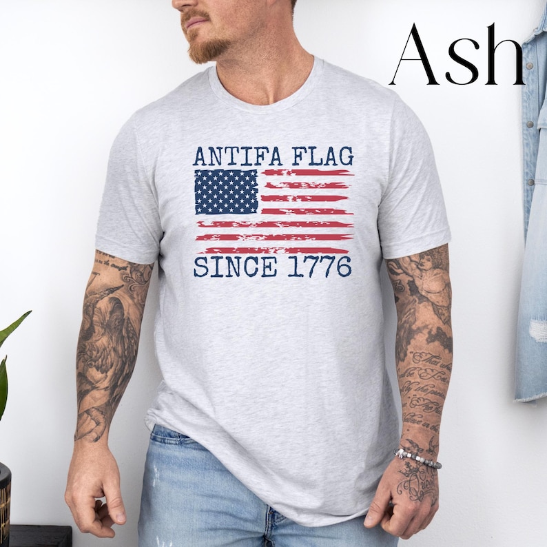 May include: Ash-colored t-shirt featuring a distressed American flag design with the text "ANTIFA FLAG SINCE 1776" in navy blue. The shirt is a crew neck style and appears to be made of a soft, comfortable material.