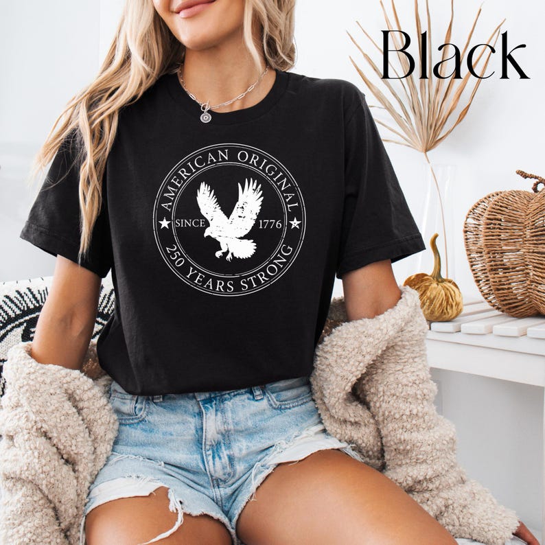 May include: Black t-shirt featuring a white circular graphic with an eagle and the text "AMERICAN ORIGINAL SINCE 1776 250 YEARS STRONG." The shirt is short-sleeved and worn with denim shorts and a cream-colored sweater.