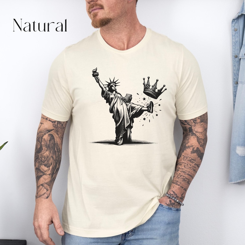 Może przedstawiać: Kremowa koszulka z napisem "Natural" u g&oacute;ry. Koszulka przedstawia czarno-białą grafikę Statuy Wolności kopiącej koronę. Statua Wolności trzyma pochodnię i książkę.