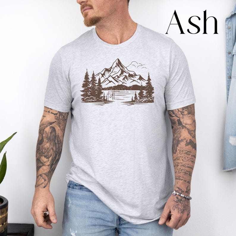 May include: A gray t-shirt with a brown mountain scene graphic. The graphic features a mountain range, a lake, and trees. The text "Ash" is visible in the upper right corner of the image.