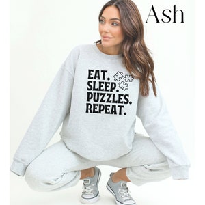 May include: A gray sweatshirt with the text "EAT. SLEEP. PUZZLES. REPEAT." and a graphic of puzzle pieces. The model is wearing the sweatshirt and gray sweatpants.