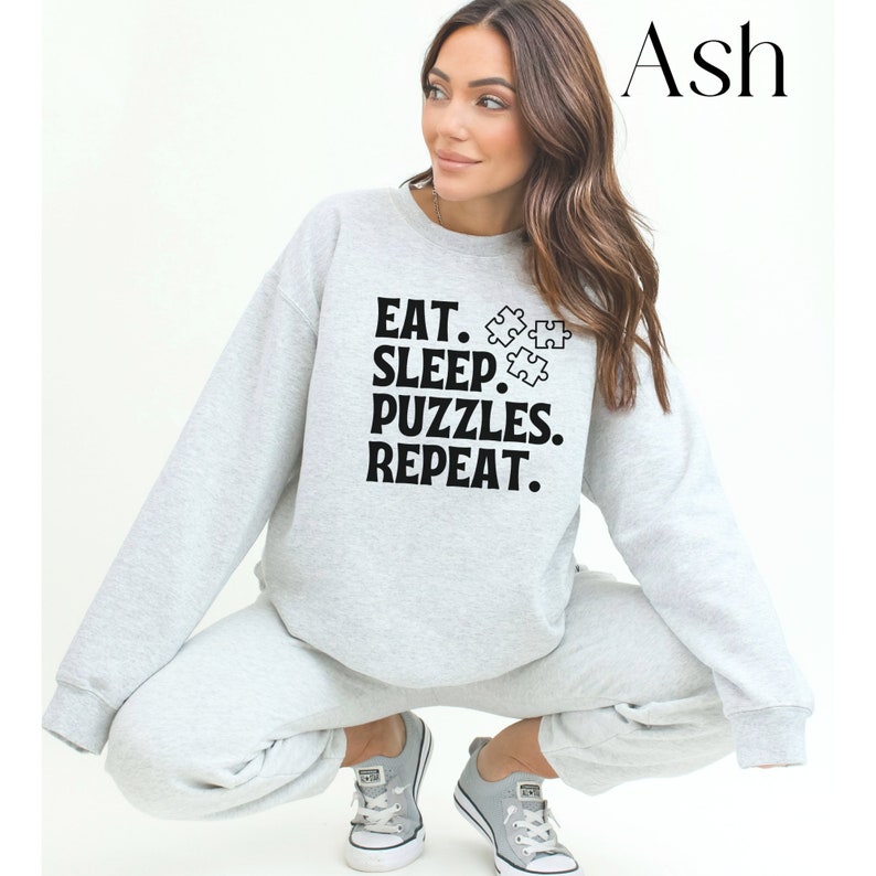 May include: A gray sweatshirt with the text "EAT. SLEEP. PUZZLES. REPEAT." and a graphic of puzzle pieces. The model is wearing the sweatshirt and gray sweatpants.