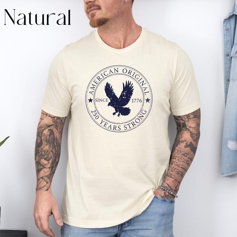 May include: Cream-colored t-shirt featuring a navy blue circular graphic with an eagle and the text "AMERICAN ORIGINAL SINCE 1776 250 YEARS STRONG". The word "Natural" is printed in black in the upper left corner.