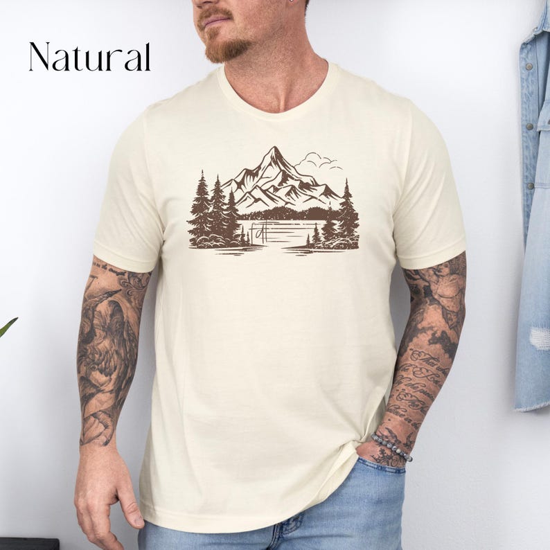 May include: A cream colored t-shirt with a brown mountain scene graphic. The graphic features a mountain range with a lake and trees.