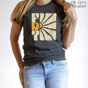 May include: A dark heather grey t-shirt with a vintage-style graphic of a sun with the word "First" in yellow letters.