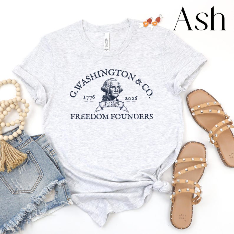 May include: Light gray t-shirt with a navy blue graphic featuring George Washington's portrait and the text "G. Washington & Co. 1776-2026 Freedom Founders." The shirt is paired with denim shorts and studded sandals.
