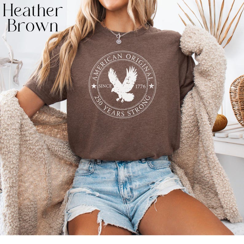 May include: Heather brown t-shirt with a circular graphic featuring an eagle and the text "AMERICAN ORIGINAL SINCE 1776 250 YEARS STRONG." The shirt is worn with denim shorts and a cream-colored cardigan.