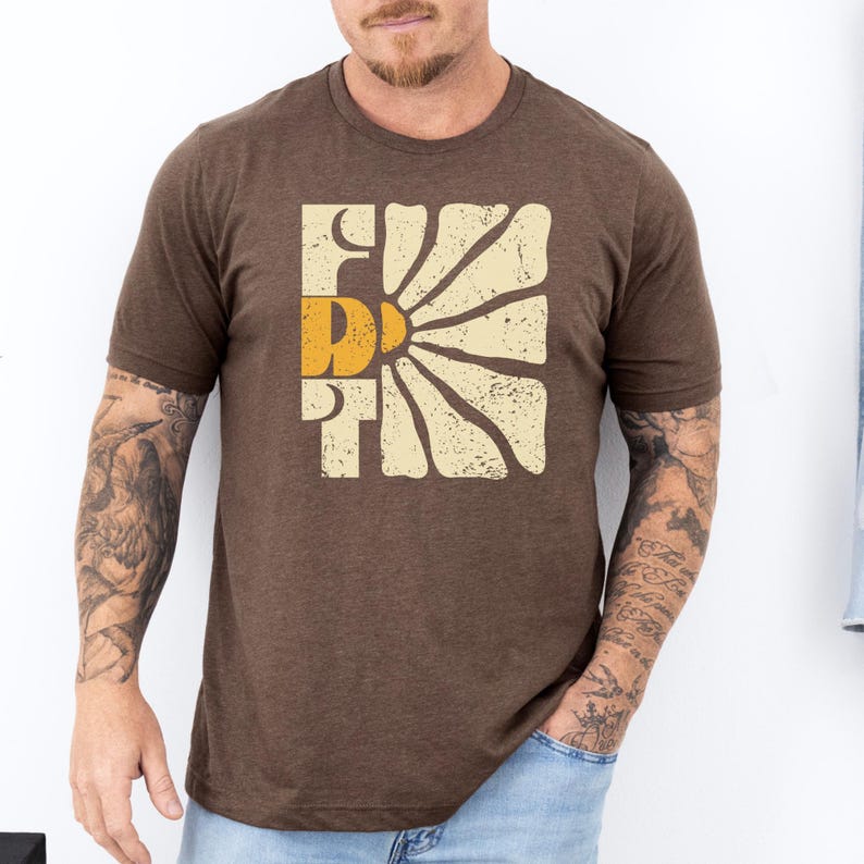 May include: A brown t-shirt featuring a vintage-style graphic of a sun rising behind the word "FIRST". The graphic is in shades of yellow, orange, and white.