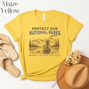 May include: Yellow t-shirt with a brown bear and a small bear cub sitting by a lake in a forest. The text "Protect Our National Parks" is printed in blue and brown on the shirt. The text "Resist. Persist. Prevail." is printed in brown below the image.