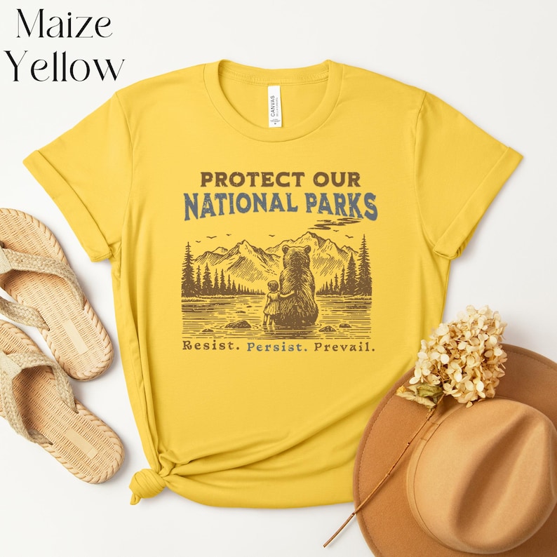 May include: Yellow t-shirt with a brown bear and a small bear cub sitting by a lake in a forest. The text "Protect Our National Parks" is printed in blue and brown on the shirt. The text "Resist. Persist. Prevail." is printed in brown below the image.