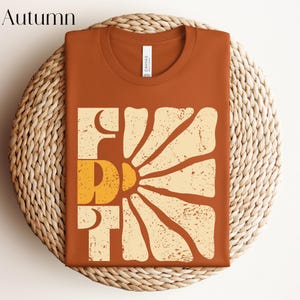 May include: A brown t-shirt featuring a vintage-style graphic of a sun with the word "First" printed in white and yellow.