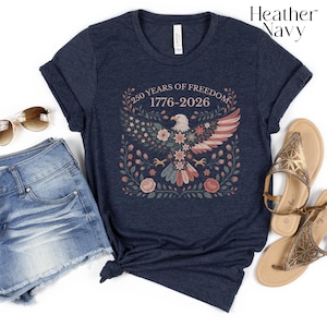 May include: Heather navy t-shirt featuring a floral design with an eagle, stars, and stripes. The text reads "250 YEARS OF FREEDOM 1776-2026". The shirt is paired with denim shorts and sandals, suggesting a casual, patriotic theme.
