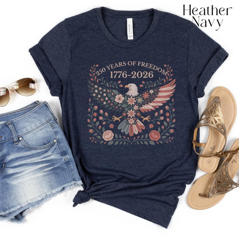 May include: Heather navy t-shirt featuring a floral design with an eagle, stars, and stripes. The text reads "250 YEARS OF FREEDOM 1776-2026". The shirt is paired with denim shorts and sandals, suggesting a casual, patriotic theme.