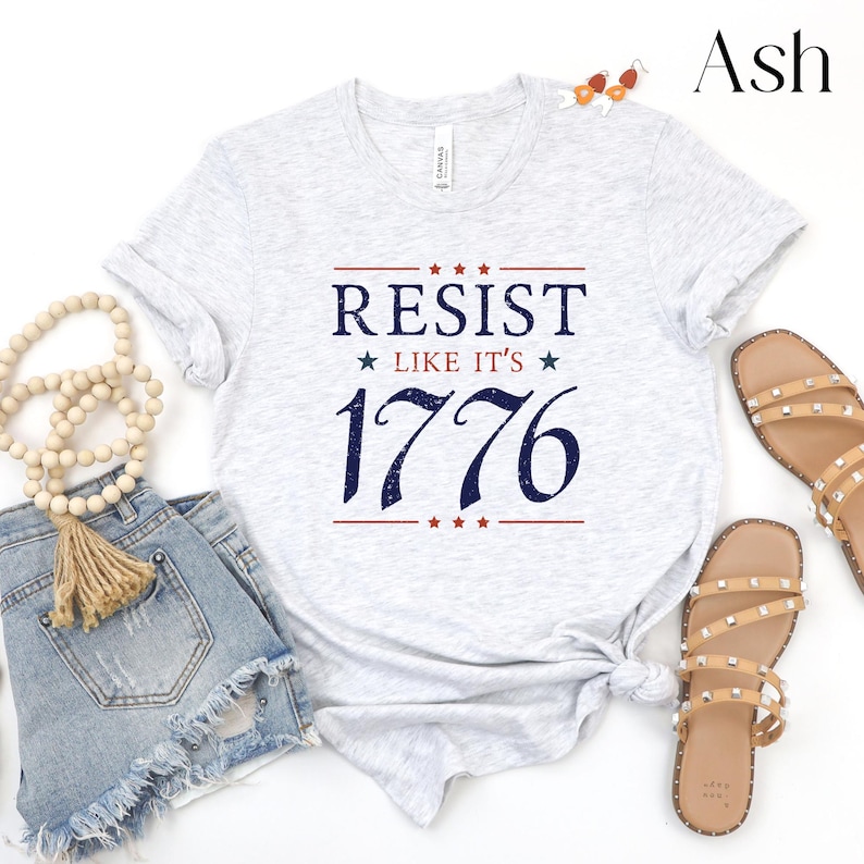 Pode incluir: Camiseta cinza claro com as palavras "RESIST LIKE IT'S 1776" em azul marinho e vermelho. A camiseta &eacute; estilizada com shorts jeans, bijuterias de contas e sand&aacute;lias com tachas. A palavra "Ash" est&aacute; no canto superior direito.