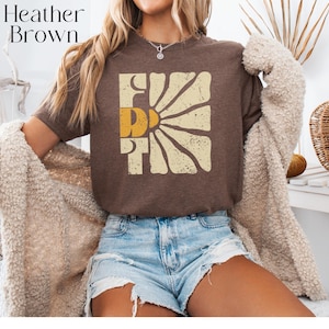 May include: A brown t-shirt with a vintage-style graphic of a sun with the word "First" in a yellow and cream colour scheme.