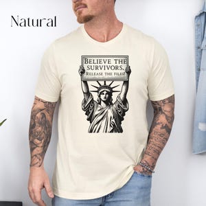 May include: Cream-coloured t-shirt with a black and white illustration of the Statue of Liberty holding a sign. The sign reads "BELIEVE THE SURVIVORS. RELEASE THE FILES!" The word "Natural" is printed in the upper left corner.