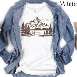 May include: White t-shirt with a brown graphic of a mountain range, lake, and trees.