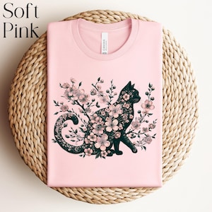 May include: A soft pink t-shirt featuring a black cat silhouette filled with pink floral blossoms. The shirt has a crew neck and the text "Soft Pink" is in the upper left corner. The shirt is displayed on a woven, circular surface.