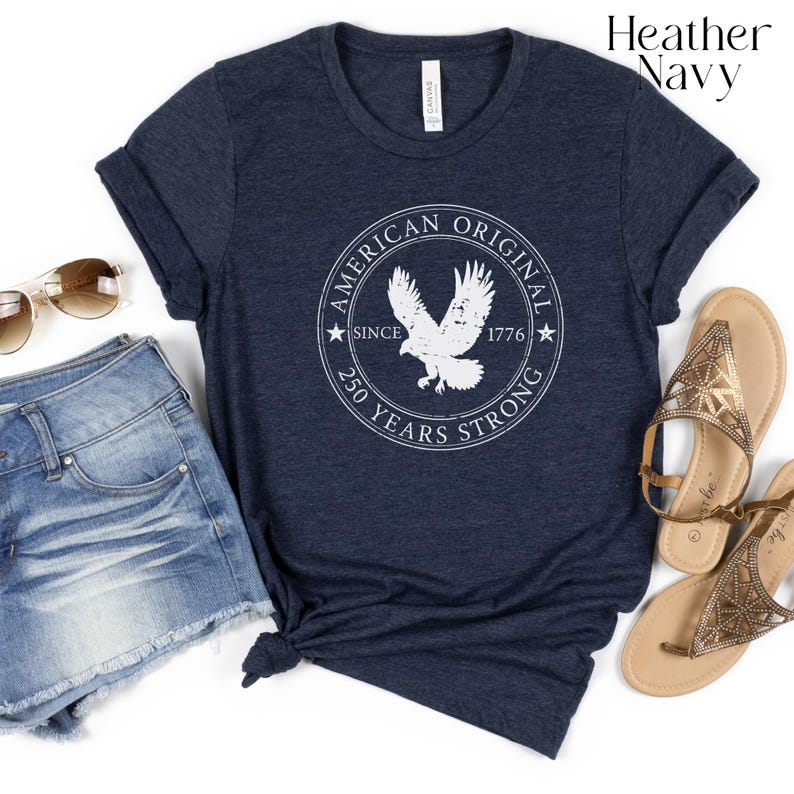 May include: Heather navy blue t-shirt with a white circular graphic featuring an eagle and the text "AMERICAN ORIGINAL SINCE 1776 250 YEARS STRONG." The shirt is paired with denim shorts and gold sandals.