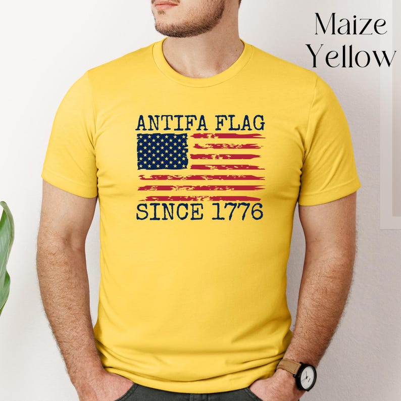 May include: Maize yellow t-shirt with the text "ANTIFA FLAG SINCE 1776" and a distressed American flag design. The flag features a blue rectangle with white stars and red and white stripes. The shirt is short-sleeved.