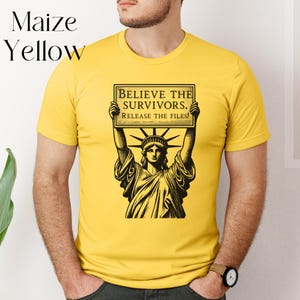 May include: Maize yellow t-shirt with a black and white graphic of the Statue of Liberty holding a sign. The sign reads "BELIEVE THE SURVIVORS. RELEASE THE FILES!". The shirt is a solid colour and has short sleeves.