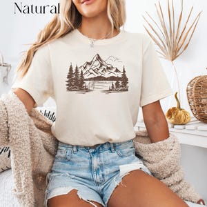 May include: A white t-shirt with a brown graphic of a mountain range, trees, and a lake. The text "Natural" is in the top left corner of the image.