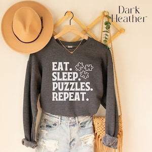 May include: Dark heather grey sweatshirt with the text "Eat. Sleep. Puzzles. Repeat." in white lettering. The sweatshirt is hanging on a wooden hanger with a brown hat and a woven bag.
