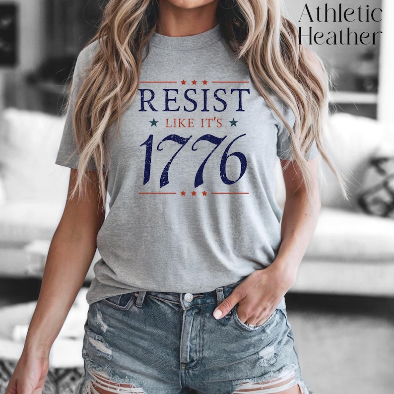 Pode incluir: T-shirt cinzento mesclado com a frase "RESIST LIKE IT'S 1776" em azul marinho e vermelho. O design apresenta estrelas e um tema patri&oacute;tico. A camisa tem gola redonda.