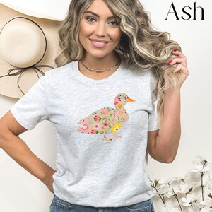 Peut inclure: Un t-shirt gris clair avec un motif de canard floral. Le canard est d&eacute;limit&eacute; par du brun et rempli de fleurs color&eacute;es.