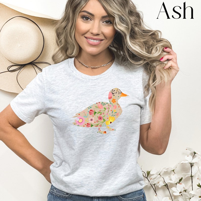 Peut inclure: Un t-shirt gris clair avec un motif de canard floral. Le canard est d&eacute;limit&eacute; par du brun et rempli de fleurs color&eacute;es.