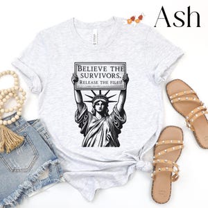 May include: Light grey t-shirt with a black and white graphic of the Statue of Liberty holding a sign that reads "BELIEVE THE SURVIVORS. RELEASE THE FILES!". The shirt is styled with denim shorts and sandals.