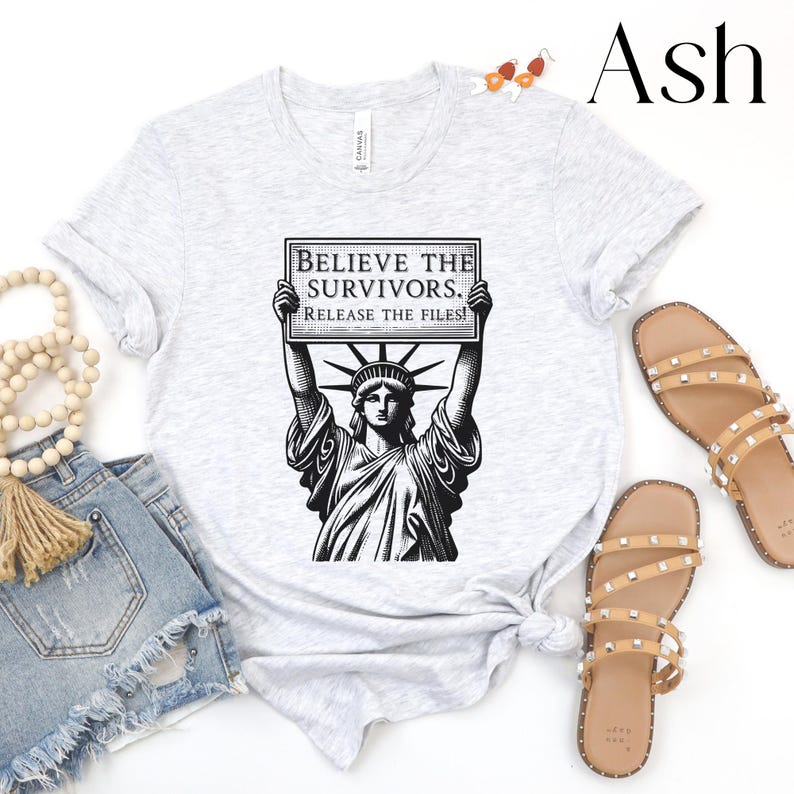 May include: Light grey t-shirt with a black and white graphic of the Statue of Liberty holding a sign that reads "BELIEVE THE SURVIVORS. RELEASE THE FILES!". The shirt is styled with denim shorts and sandals.