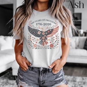 May include: Ash-colored t-shirt featuring a detailed graphic of an eagle with a floral design and the text "250 YEARS OF FREEDOM 1776-2026". The eagle's wings are colored in the style of the American flag. The design is surrounded by floral elements.