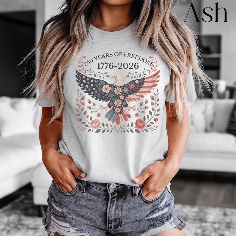 May include: Ash-colored t-shirt featuring a detailed graphic of an eagle with a floral design and the text "250 YEARS OF FREEDOM 1776-2026". The eagle's wings are colored in the style of the American flag. The design is surrounded by floral elements.