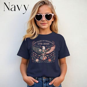 May include: Navy blue t-shirt featuring a patriotic design with an eagle, floral elements, and the text "250 YEARS OF FREEDOM 1776-2026". The shirt is worn by a person wearing heart-shaped sunglasses.