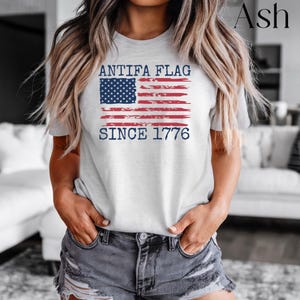 May include: Ash-colored t-shirt featuring a distressed American flag design with the text "ANTIFA FLAG SINCE 1776" in navy blue. The shirt is a crew neck and is made of a soft material.