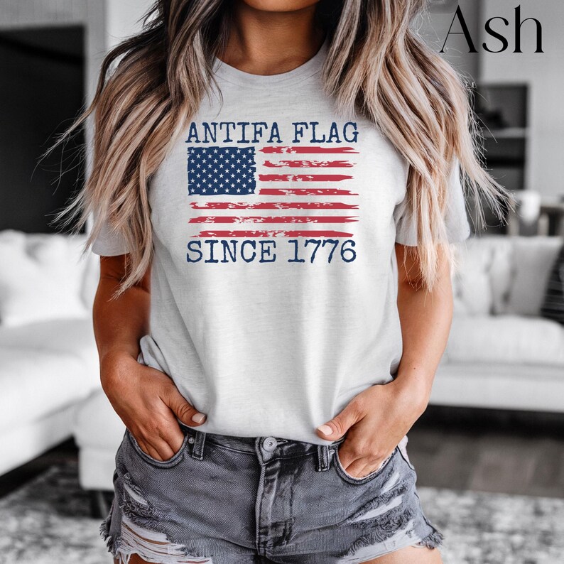 May include: Ash-colored t-shirt featuring a distressed American flag design with the text "ANTIFA FLAG SINCE 1776" in navy blue. The shirt is a crew neck and is made of a soft material.