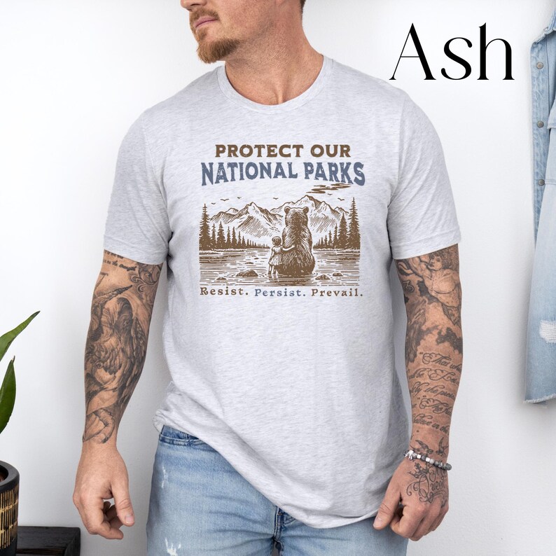 May include: A light grey t-shirt with a brown bear and cub illustration. The text "Protect Our National Parks" is printed in brown on the shirt. The text "Resist. Persist. Prevail." is printed in blue below the illustration.