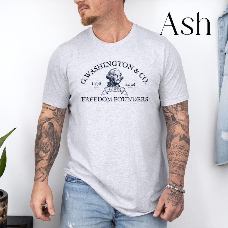 May include: Ash-colored t-shirt with a navy blue graphic. The graphic reads "G. Washington & Co. 1776-2026 Freedom Founders" with a portrait of George Washington. The shirt is short-sleeved and made of a soft material.
