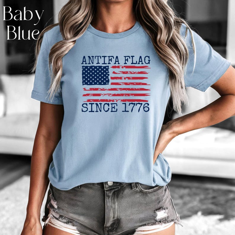 May include: Baby blue t-shirt with the text "ANTIFA FLAG SINCE 1776" and a distressed American flag design in red, white, and blue. The shirt is short-sleeved and made of a soft material.