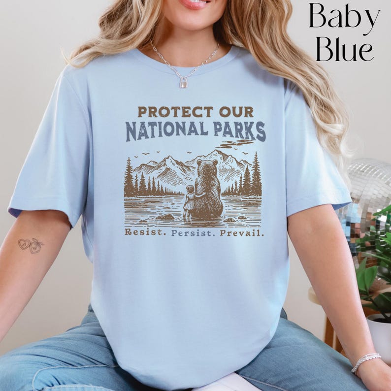 May include: A light blue t-shirt with a brown bear and a small bear cub sitting in front of a mountain scene. The text "PROTECT OUR NATIONAL PARKS" is printed in dark blue above the image. The text "Resist. Persist. Prevail." is printed in brown below the image.