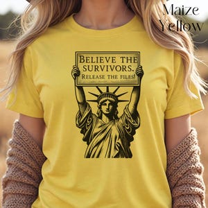 May include: Maize yellow t-shirt with a black graphic of the Statue of Liberty holding a sign. The sign reads "BELIEVE THE SURVIVORS. RELEASE THE FILES!" The shirt is a short-sleeved crew neck.