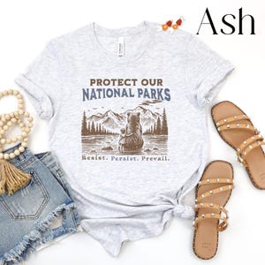 May include: A light gray t-shirt with the text "Protect Our National Parks" in blue and brown lettering. A brown bear and a smaller bear cub are sitting on a rock by a lake with mountains and trees in the background. The text "Resist. Persist. Prevail." is printed below the image.