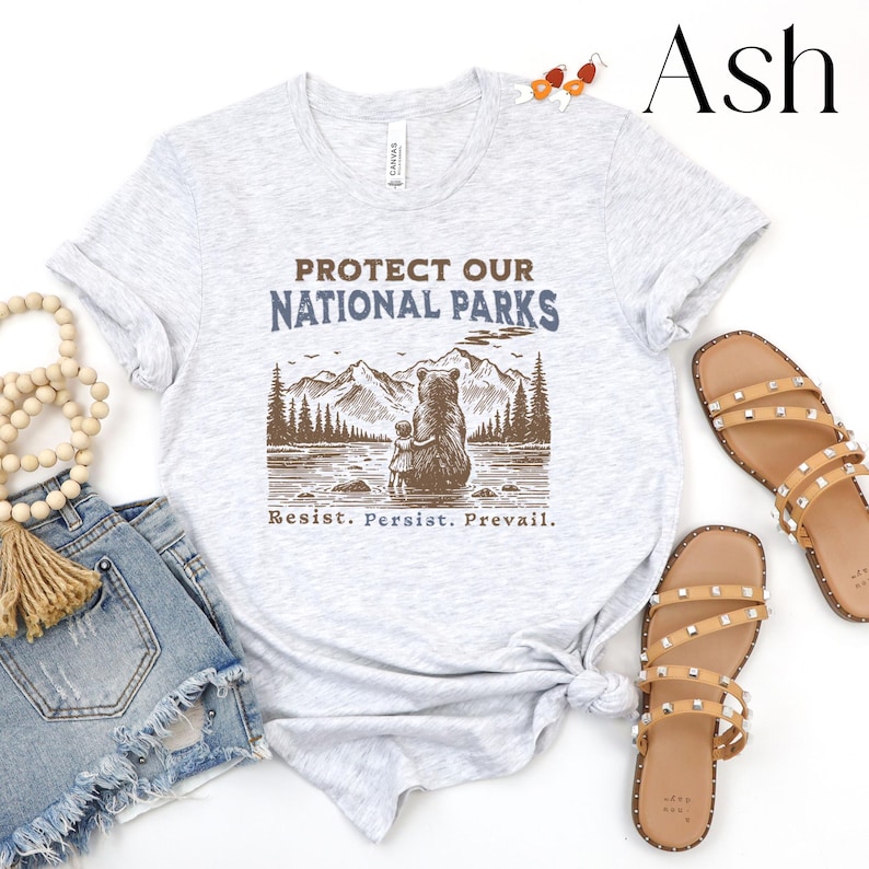 May include: A light gray t-shirt with the text "Protect Our National Parks" in blue and brown lettering. A brown bear and a smaller bear cub are sitting on a rock by a lake with mountains and trees in the background. The text "Resist. Persist. Prevail." is printed below the image.