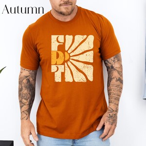 May include: A rust-coloured t-shirt with a retro sunburst graphic and the word "Friday" in a vintage font.