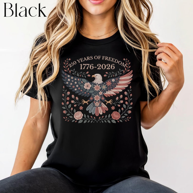 May include: Black t-shirt featuring a patriotic design. The graphic includes an eagle with wings in the colors of the American flag, surrounded by floral elements. The text "250 YEARS OF FREEDOM 1776-2026" is arched above the eagle.