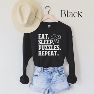 May include: Black sweatshirt with white text that reads "Eat. Sleep. Puzzles. Repeat." and a graphic of three puzzle pieces.