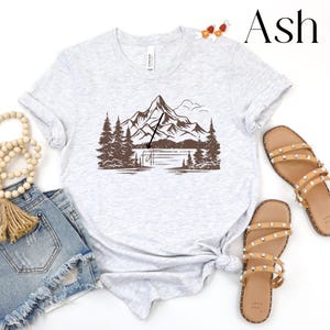 May include: A gray t-shirt with a brown mountain and lake graphic. The graphic features a mountain range with a lake in the foreground and a fishing rod in the air. The text "Ash" is in the upper right corner of the image.